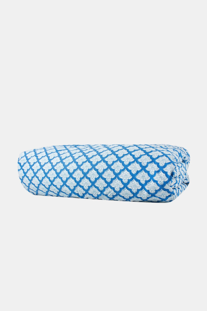 A neatly made bed features the Roller Rabbit Jemina Duvet Cover in blue and white, set before a white curtain. A small bedside table holds a striped vase with flowers, books, and a lamp atop a woven rug.