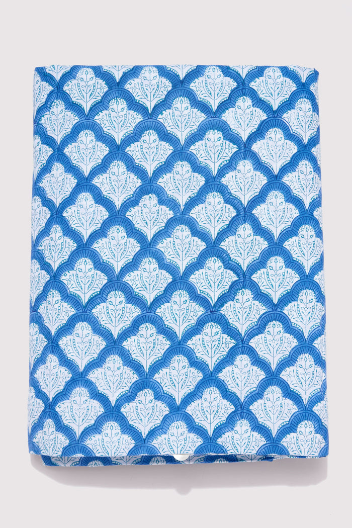 A neatly made bed features the Roller Rabbit Jemina Duvet Cover in blue and white, set before a white curtain. A small bedside table holds a striped vase with flowers, books, and a lamp atop a woven rug.