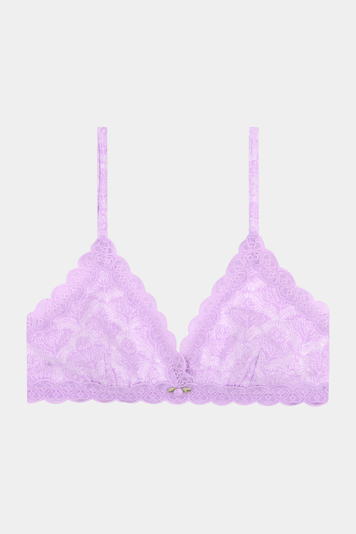 A woman with wavy brown hair poses confidently in a matching light purple scalloped lace trim Roller Rabbit Jemina Jane Bralette and underwear set against a plain, light background.