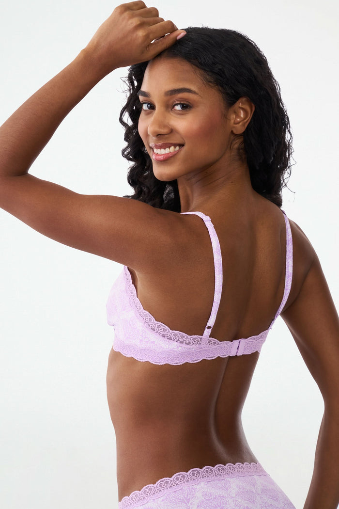 A woman with wavy brown hair poses confidently in a matching light purple scalloped lace trim Roller Rabbit Jemina Jane Bralette and underwear set against a plain, light background.