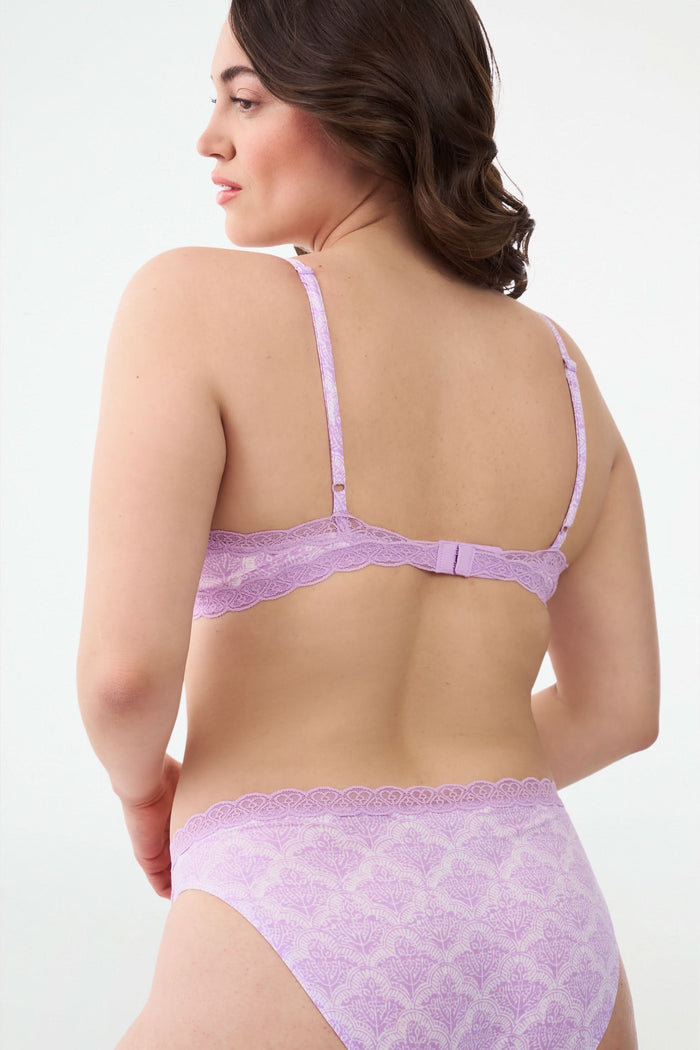 A woman with wavy brown hair poses confidently in a matching light purple scalloped lace trim Roller Rabbit Jemina Jane Bralette and underwear set against a plain, light background.