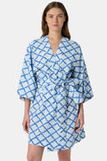 A woman stands barefoot against a white background, wearing the Roller Rabbit Jemina Kimono—a blue and white cotton piece featuring a Jemina print, wide sleeves, and a tied belt.