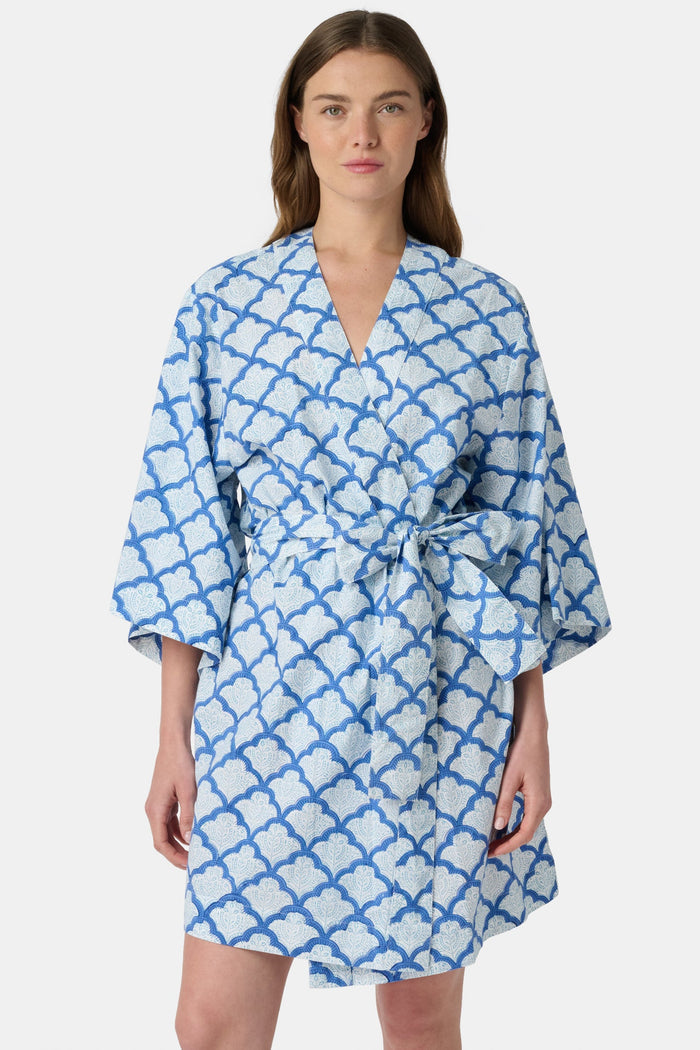 A woman stands barefoot against a white background, wearing the Roller Rabbit Jemina Kimono—a blue and white cotton piece featuring a Jemina print, wide sleeves, and a tied belt.