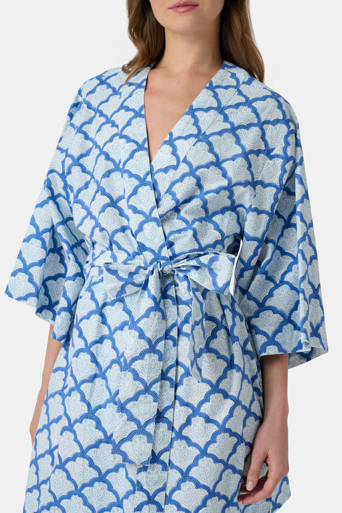 A woman stands barefoot against a white background, wearing the Roller Rabbit Jemina Kimono—a blue and white cotton piece featuring a Jemina print, wide sleeves, and a tied belt.
