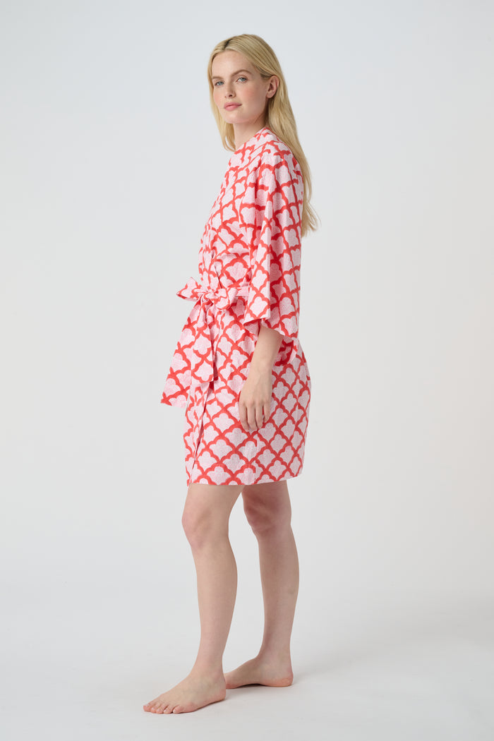 A woman stands barefoot in the Roller Rabbit Jemina Kimono, a knee-length cotton design with wide sleeves, a belt, and a red and white Jemina print. She faces forward against a plain light background.