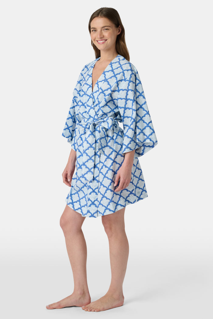 A woman stands barefoot against a white background, wearing the Roller Rabbit Jemina Kimono—a blue and white cotton piece featuring a Jemina print, wide sleeves, and a tied belt.