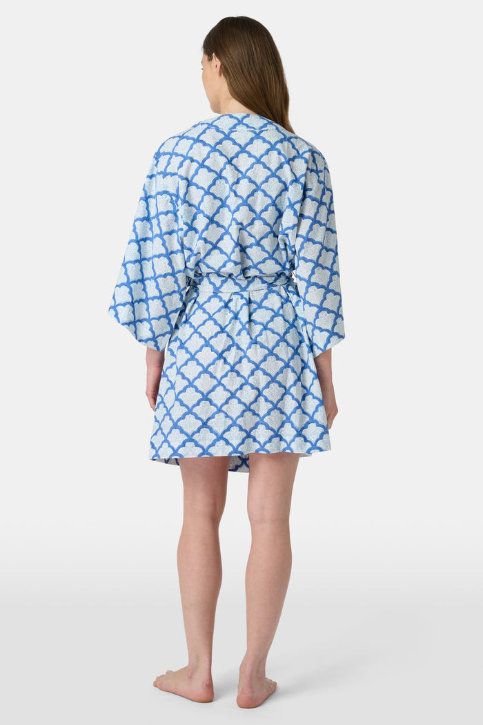 A woman stands barefoot against a white background, wearing the Roller Rabbit Jemina Kimono—a blue and white cotton piece featuring a Jemina print, wide sleeves, and a tied belt.