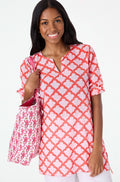A woman with long dark hair smiles in a relaxed fit, pink and red Jemina Kurta by Roller Rabbit, holding a matching white and pink patterned tote bag against a plain light background.