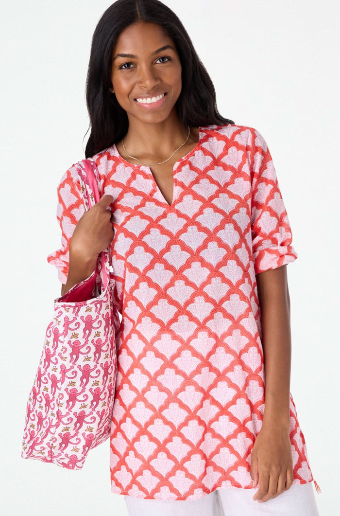 A woman with long dark hair smiles in a relaxed fit, pink and red Jemina Kurta by Roller Rabbit, holding a matching white and pink patterned tote bag against a plain light background.