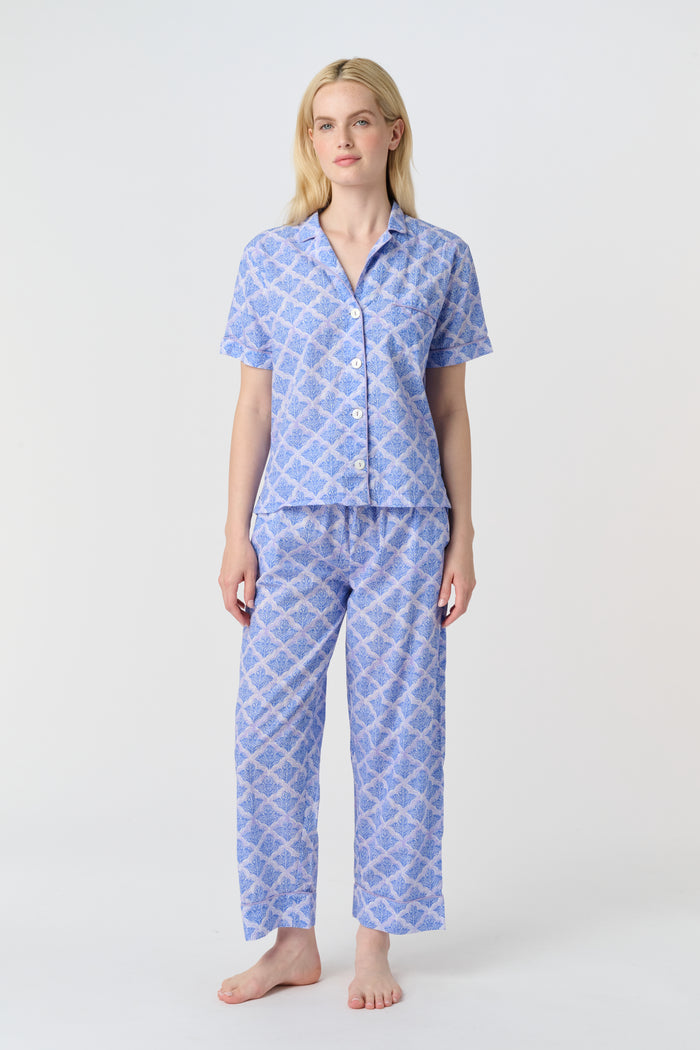 A person stands barefoot against a plain background, wearing the Roller Rabbit Jemina Liza Set—a light blue cotton voile loungewear set with a geometric pattern, featuring a short-sleeve button-up shirt and loose-fitting pants.