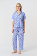 A person stands barefoot against a plain background, wearing the Roller Rabbit Jemina Liza Set—a light blue cotton voile loungewear set with a geometric pattern, featuring a short-sleeve button-up shirt and loose-fitting pants.