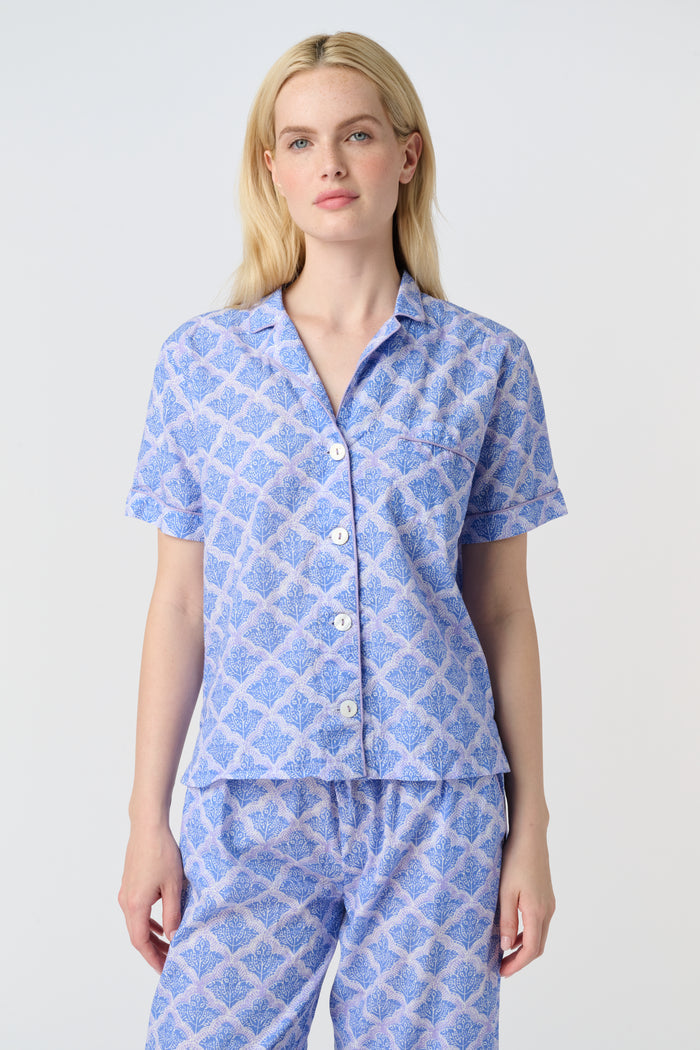 A person stands barefoot against a plain background, wearing the Roller Rabbit Jemina Liza Set—a light blue cotton voile loungewear set with a geometric pattern, featuring a short-sleeve button-up shirt and loose-fitting pants.