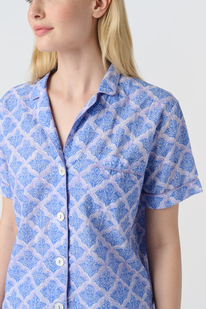 A person stands barefoot against a plain background, wearing the Roller Rabbit Jemina Liza Set—a light blue cotton voile loungewear set with a geometric pattern, featuring a short-sleeve button-up shirt and loose-fitting pants.
