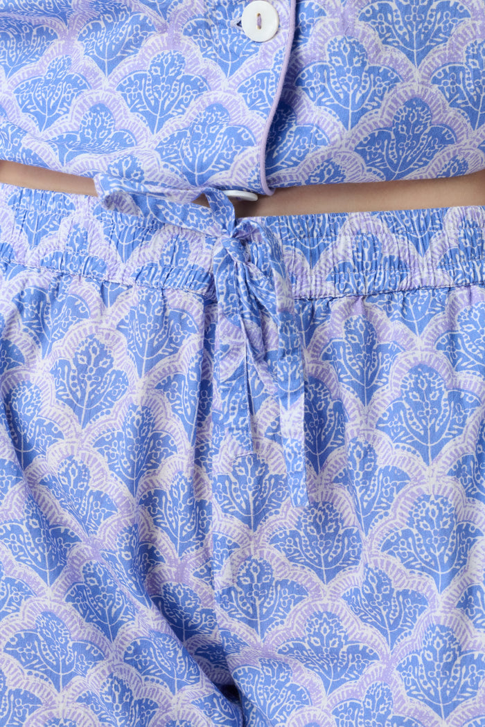 A person stands barefoot against a plain background, wearing the Roller Rabbit Jemina Liza Set—a light blue cotton voile loungewear set with a geometric pattern, featuring a short-sleeve button-up shirt and loose-fitting pants.