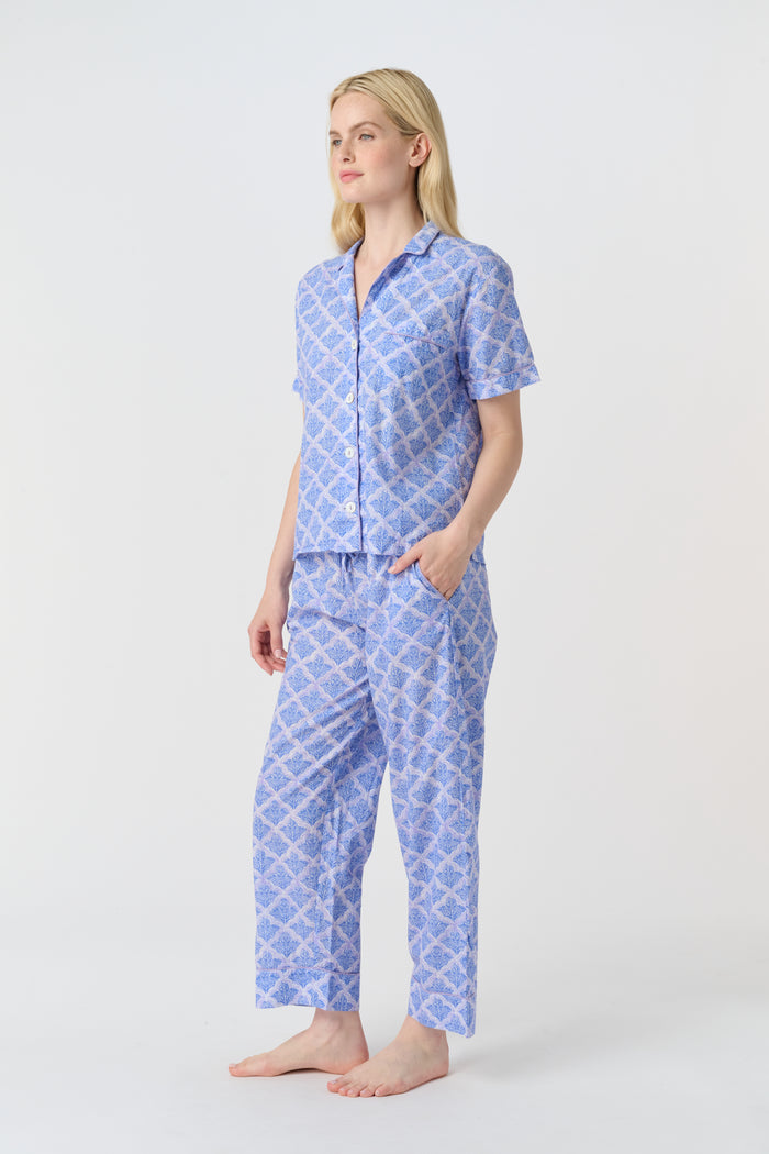 A person stands barefoot against a plain background, wearing the Roller Rabbit Jemina Liza Set—a light blue cotton voile loungewear set with a geometric pattern, featuring a short-sleeve button-up shirt and loose-fitting pants.