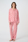 A person stands barefoot against a plain background, smiling softly and wearing the Roller Rabbit Jemina Loungewear set—a cotton silk pajama top with buttons and collar, plus loose-fitting pants—in a red and white medallion print.