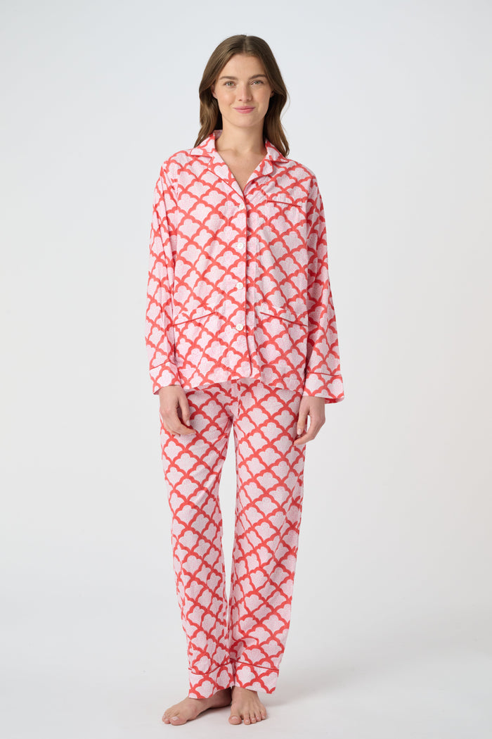 A person stands barefoot against a plain background, smiling softly and wearing the Roller Rabbit Jemina Loungewear set—a cotton silk pajama top with buttons and collar, plus loose-fitting pants—in a red and white medallion print.