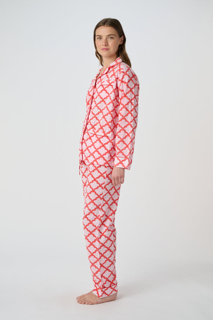 A person stands barefoot against a plain background, smiling softly and wearing the Roller Rabbit Jemina Loungewear set—a cotton silk pajama top with buttons and collar, plus loose-fitting pants—in a red and white medallion print.