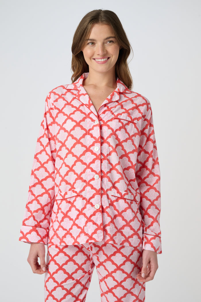 A person stands barefoot against a plain background, smiling softly and wearing the Roller Rabbit Jemina Loungewear set—a cotton silk pajama top with buttons and collar, plus loose-fitting pants—in a red and white medallion print.