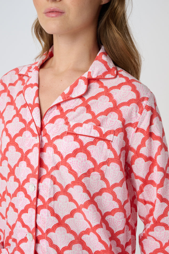 A person stands barefoot against a plain background, smiling softly and wearing the Roller Rabbit Jemina Loungewear set—a cotton silk pajama top with buttons and collar, plus loose-fitting pants—in a red and white medallion print.