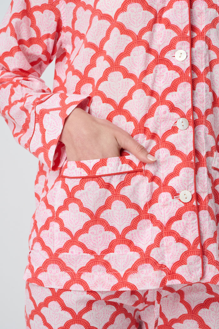 A person stands barefoot against a plain background, smiling softly and wearing the Roller Rabbit Jemina Loungewear set—a cotton silk pajama top with buttons and collar, plus loose-fitting pants—in a red and white medallion print.