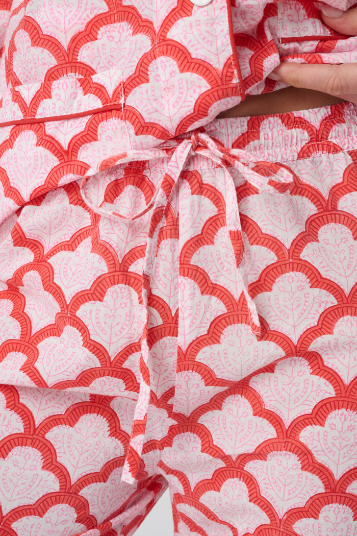 A person stands barefoot against a plain background, smiling softly and wearing the Roller Rabbit Jemina Loungewear set—a cotton silk pajama top with buttons and collar, plus loose-fitting pants—in a red and white medallion print.