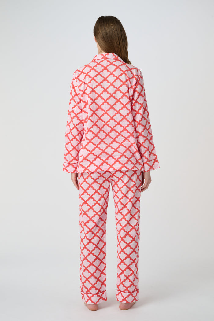 A person stands barefoot against a plain background, smiling softly and wearing the Roller Rabbit Jemina Loungewear set—a cotton silk pajama top with buttons and collar, plus loose-fitting pants—in a red and white medallion print.
