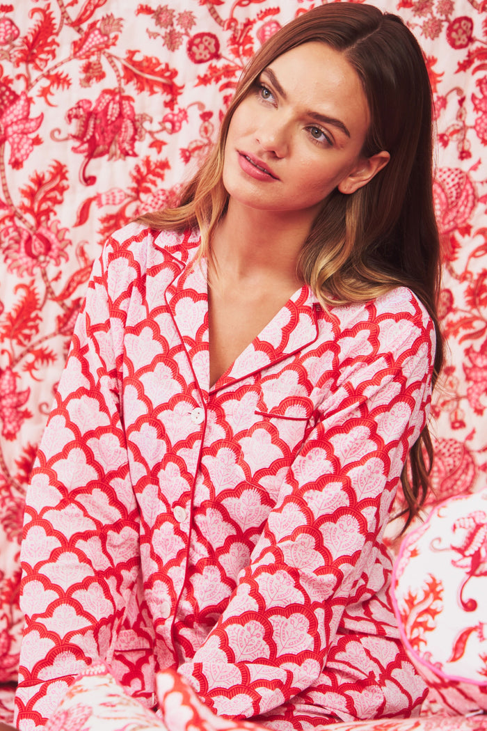 A person stands barefoot against a plain background, smiling softly and wearing the Roller Rabbit Jemina Loungewear set—a cotton silk pajama top with buttons and collar, plus loose-fitting pants—in a red and white medallion print.