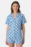 A woman wears the Roller Rabbit Jemina Lulu Set, a cotton pajama set with a matching short-sleeve button-up top and shorts, featuring a blue and white geometric leaf print, standing against a plain light background.