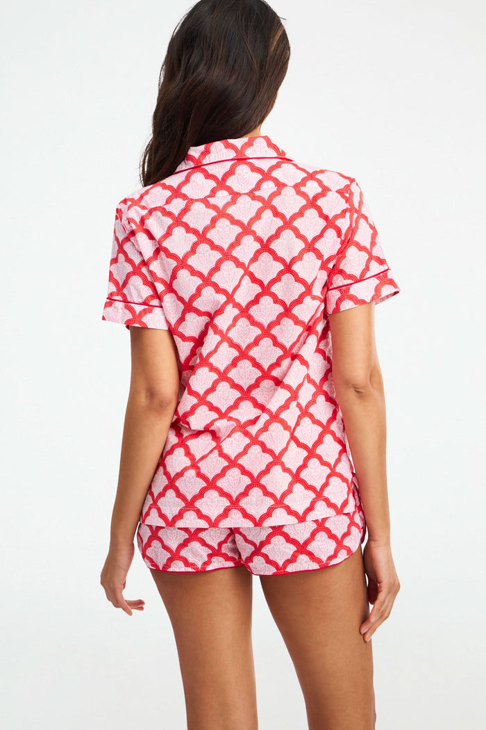 A woman with long, wavy dark hair and a relaxed expression wears the Roller Rabbit Jemina Lulu Set— a women’s pajama featuring a red and white scalloped pattern, short sleeves, and shorts—against a plain background.