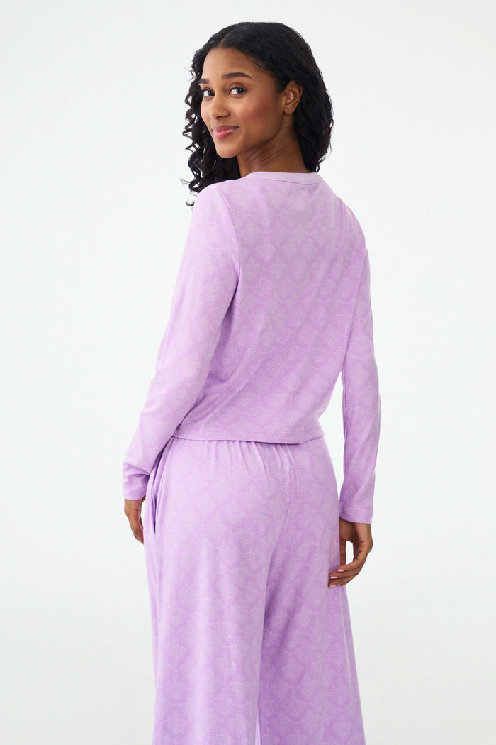 A smiling woman with curly hair wears the Roller Rabbit Jemina Nessa Cardigan pajama set in light purple, standing against a plain white background.