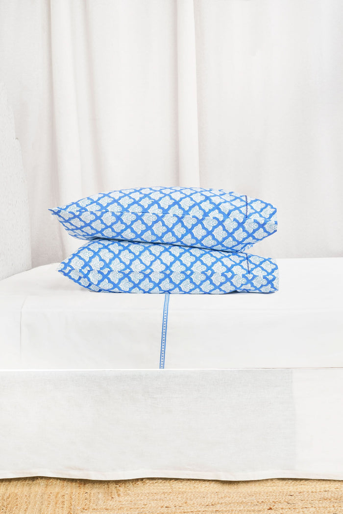 The Roller Rabbit Jemina Pillowcase Set features a blue and white geometric print, stacked on a matching bedspread with a white headboard and curtain. Made from 100 cotton, it brings style to any bedroom.