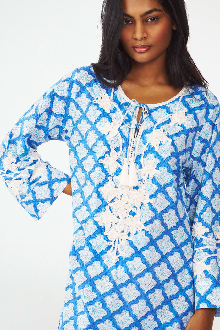 A woman stands facing forward with one hand on her hip, wearing the Roller Rabbit Jemina Punta Long Kurta in blue and white with white floral embroidery and tassel accents at the neckline.