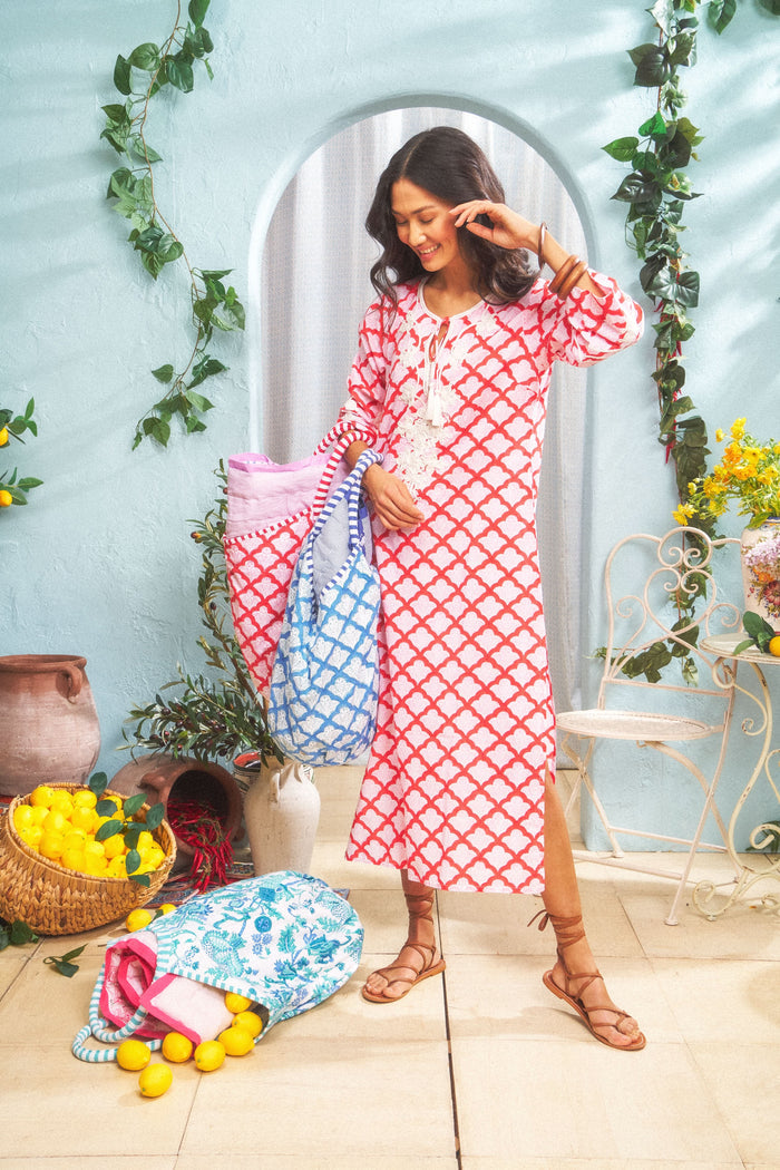 A smiling woman wears the Jemina Punta Long Kurta by Roller Rabbit, standing indoors with colorful bags. She is surrounded by lemons, baskets, greenery, and a light blue wall with a decorative arch in the background.