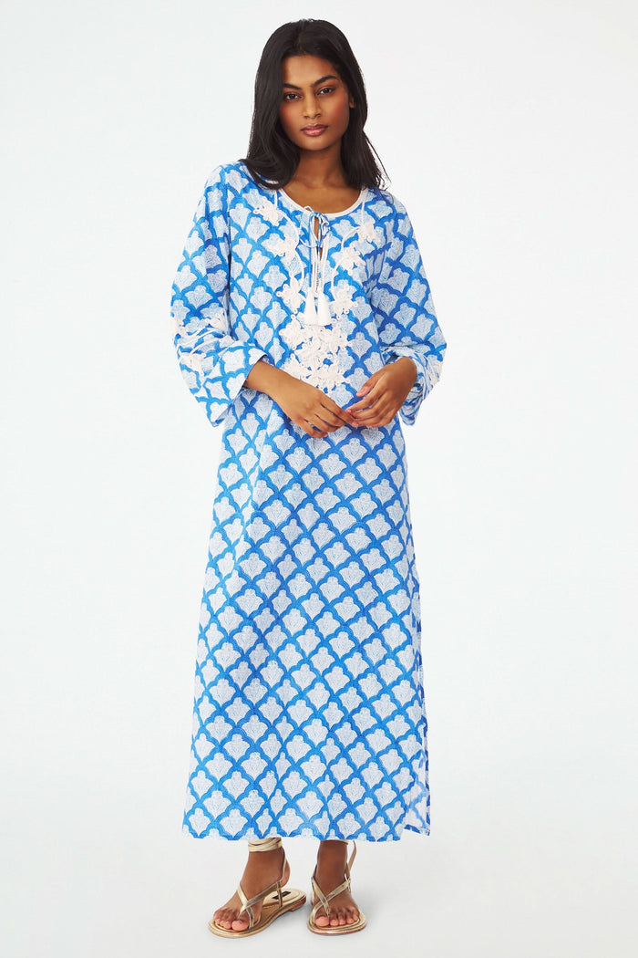 A woman stands facing forward with one hand on her hip, wearing the Roller Rabbit Jemina Punta Long Kurta in blue and white with white floral embroidery and tassel accents at the neckline.