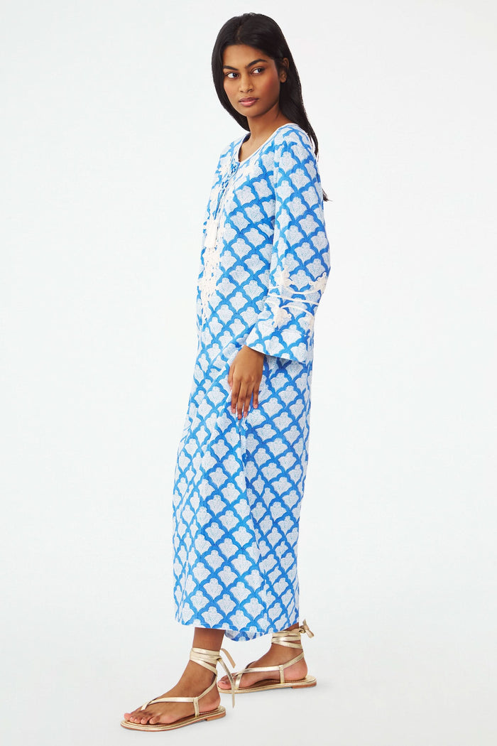 A woman stands facing forward with one hand on her hip, wearing the Roller Rabbit Jemina Punta Long Kurta in blue and white with white floral embroidery and tassel accents at the neckline.