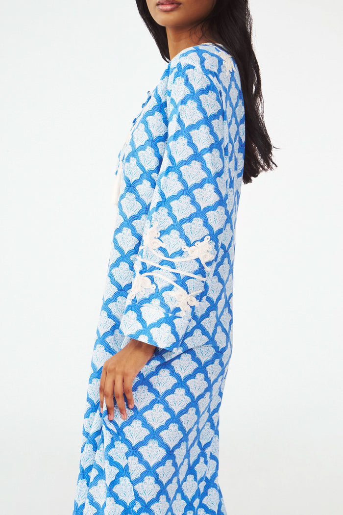 A woman stands facing forward with one hand on her hip, wearing the Roller Rabbit Jemina Punta Long Kurta in blue and white with white floral embroidery and tassel accents at the neckline.