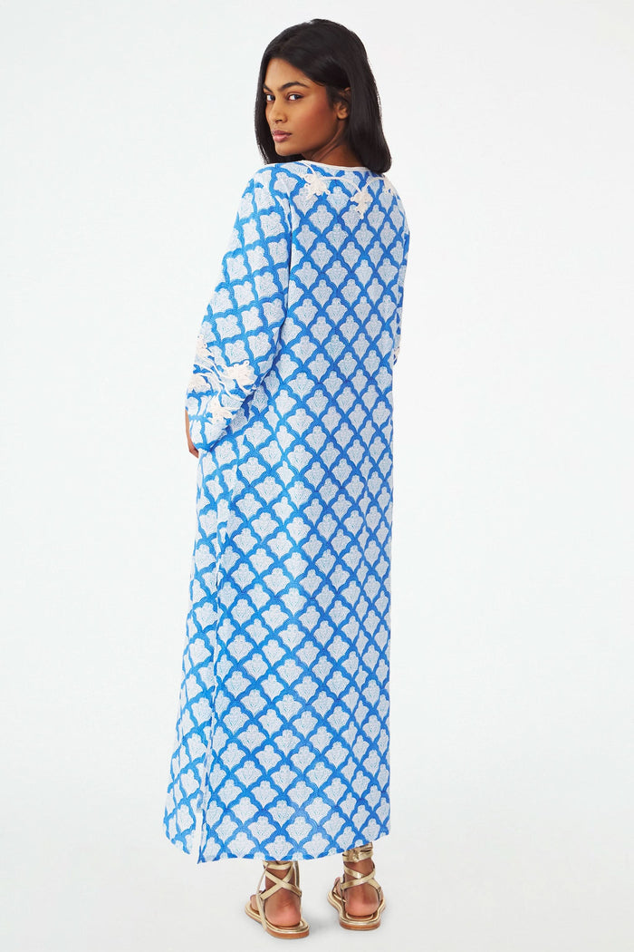 A woman stands facing forward with one hand on her hip, wearing the Roller Rabbit Jemina Punta Long Kurta in blue and white with white floral embroidery and tassel accents at the neckline.