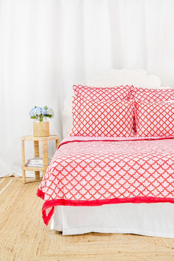 A neatly made bed features the Roller Rabbit Jemina Quilt and matching pillows, all in 100 cotton, next to a small wooden nightstand with a woven basket holding blue flowers, set against white curtains.