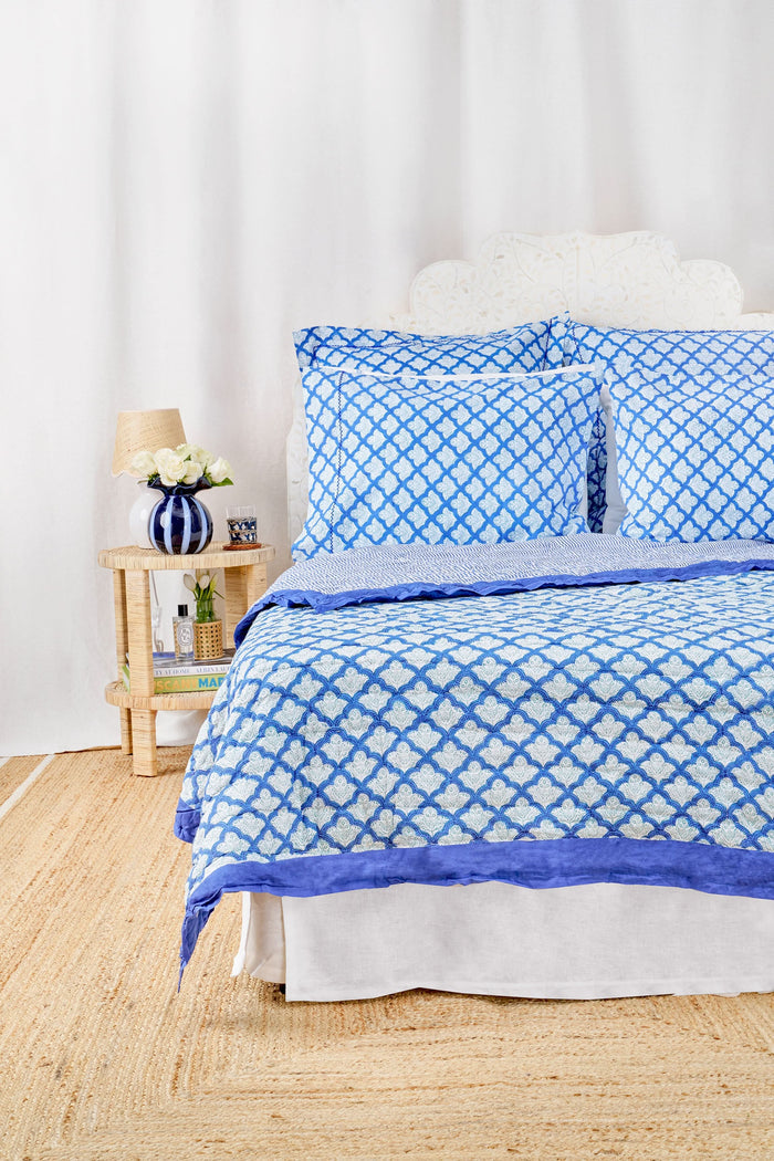 A neatly made bed features the Jemina Quilt by Roller Rabbit in blue and white, paired with pillows. It sits on a woven rug next to a wooden nightstand with a lamp, books, and flowers. White curtains hang in the background.