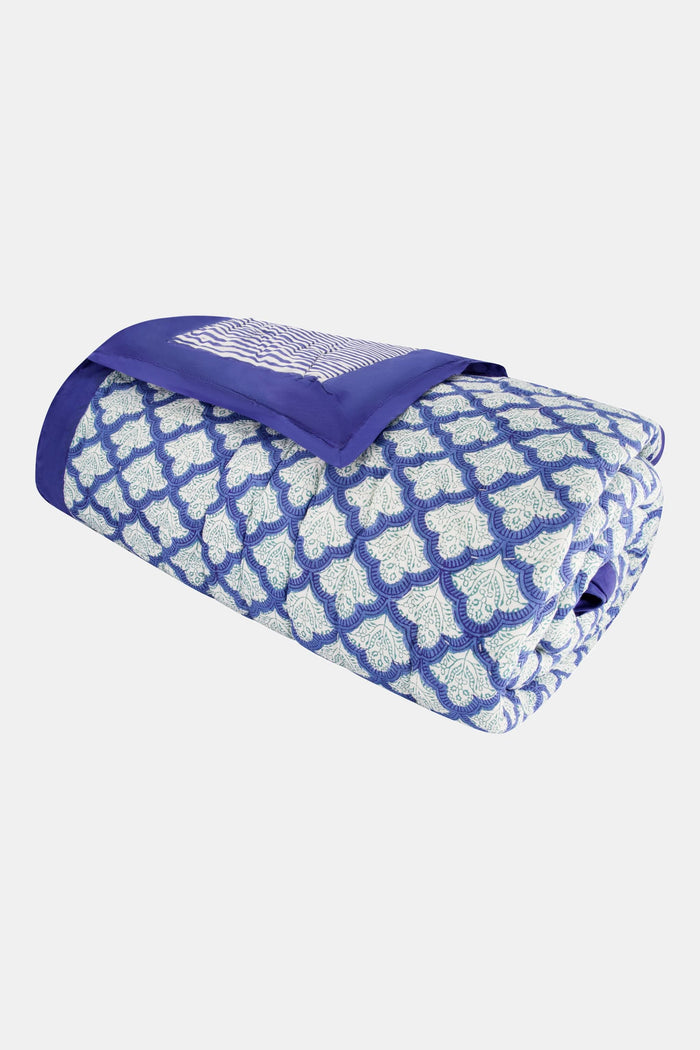 A neatly made bed features the Jemina Quilt by Roller Rabbit in blue and white, paired with pillows. It sits on a woven rug next to a wooden nightstand with a lamp, books, and flowers. White curtains hang in the background.