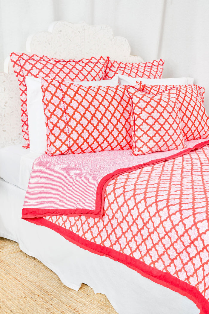 A neatly made bed features the Roller Rabbit Jemina Quilt and matching pillows, all in 100 cotton, next to a small wooden nightstand with a woven basket holding blue flowers, set against white curtains.