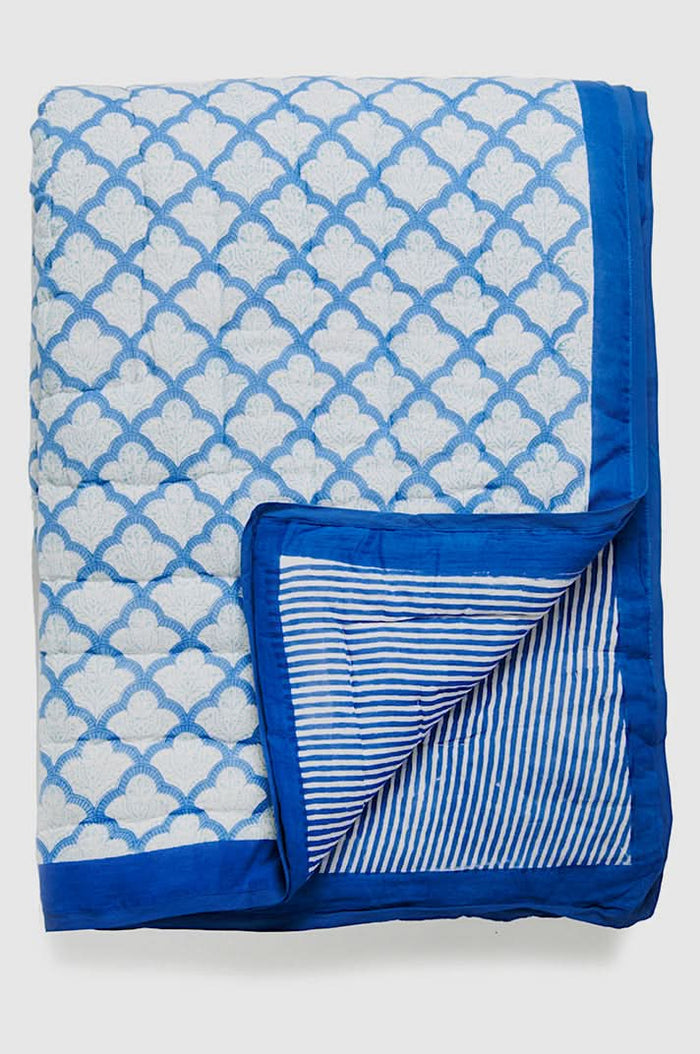 A neatly made bed features the Jemina Quilt by Roller Rabbit in blue and white, paired with pillows. It sits on a woven rug next to a wooden nightstand with a lamp, books, and flowers. White curtains hang in the background.