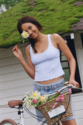 A smiling woman in the Roller Rabbit Jemina Rib Astor Tank and denim shorts stands with a bicycle. She holds a flower near her face, with a basket of colorful flowers, against a white wall and mossy roof backdrop.