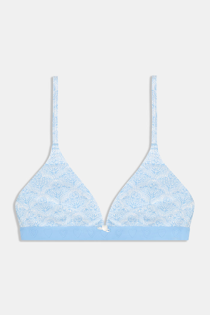 A woman with long brown hair smiles in a pale blue patterned Jemina Rib York Bralette and matching pajama shorts by Roller Rabbit, standing against a plain white background.