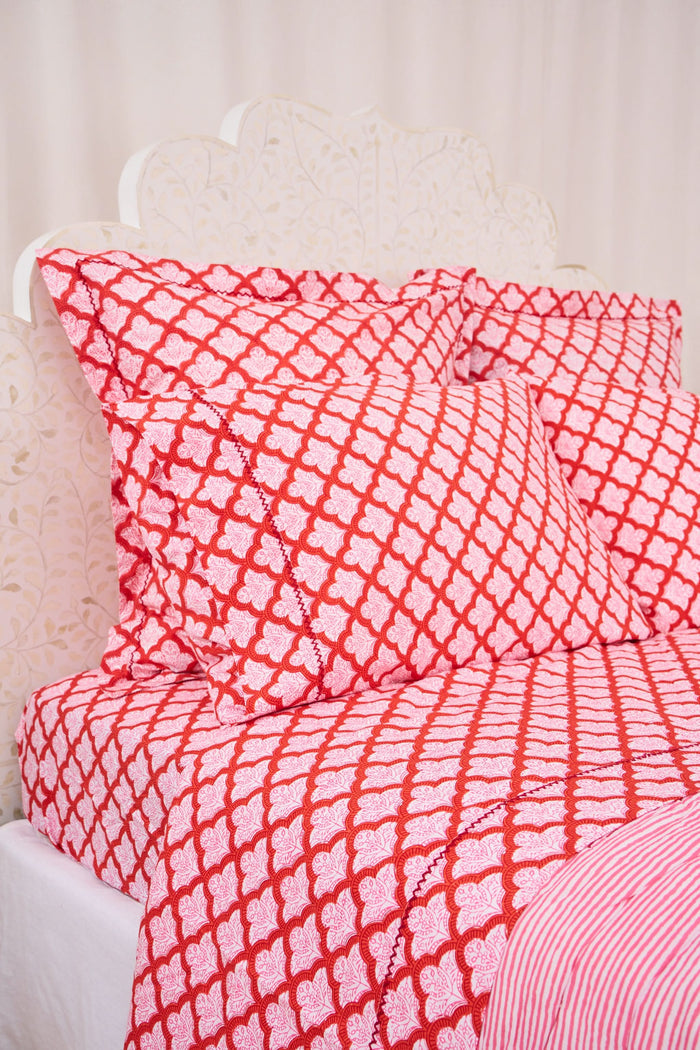 A bed with a white carved headboard showcases red and white patterned pillows and the Jemina Sheet Set by Roller Rabbit, featuring diamond and heart motifs. A pink and white striped blanket is partially visible at the edge.