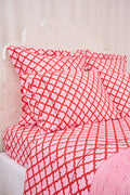 A bed with a white carved headboard showcases red and white patterned pillows and the Jemina Sheet Set by Roller Rabbit, featuring diamond and heart motifs. A pink and white striped blanket is partially visible at the edge.