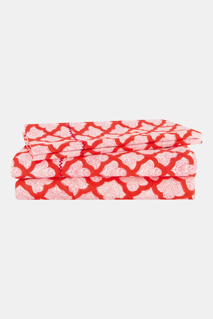 A bed with a white carved headboard showcases red and white patterned pillows and the Jemina Sheet Set by Roller Rabbit, featuring diamond and heart motifs. A pink and white striped blanket is partially visible at the edge.