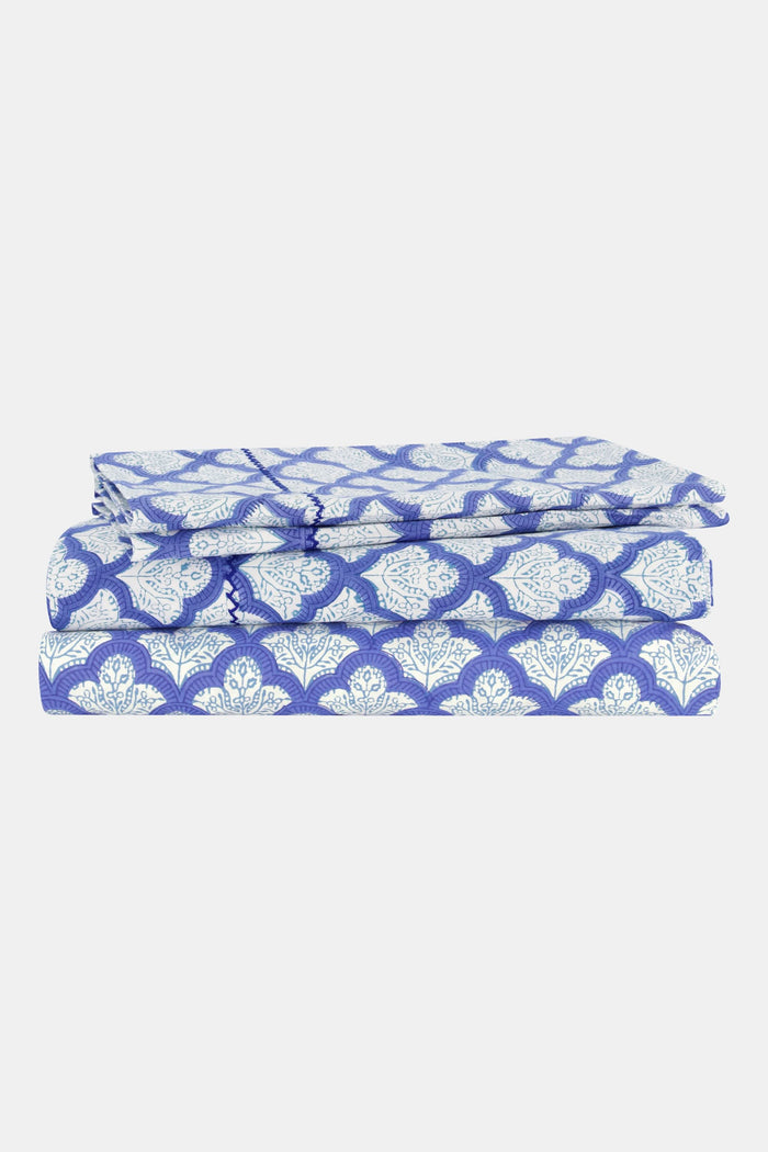 Close-up of a bed with blue and white patterned pillows and matching block-printed cotton sheets from the Jemina Sheet Set by Roller Rabbit, featuring an intricate geometric design; the headboard has a subtle white floral pattern.