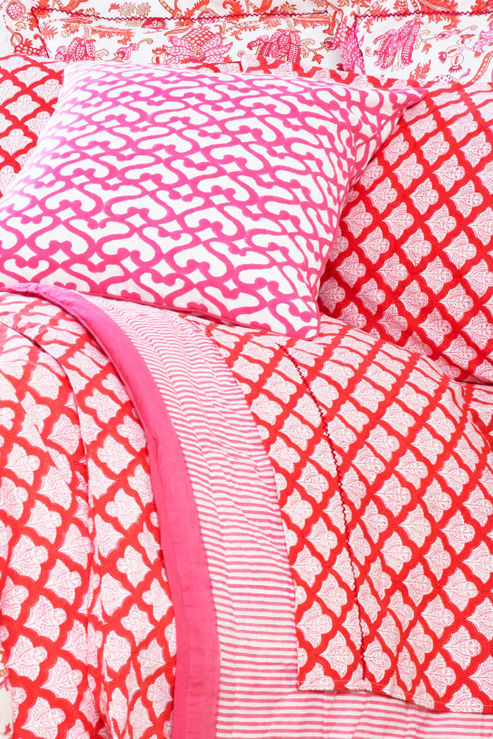 A bed with a white carved headboard showcases red and white patterned pillows and the Jemina Sheet Set by Roller Rabbit, featuring diamond and heart motifs. A pink and white striped blanket is partially visible at the edge.